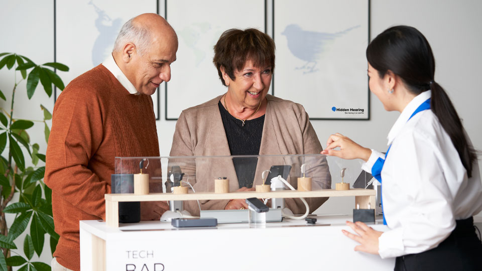 Hearing care specialist explaining different hearing aid options to two customers at a tech bar.