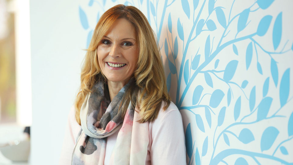 Hidden Hearing customer Sandra Pierce standing against a light blue patterned wall