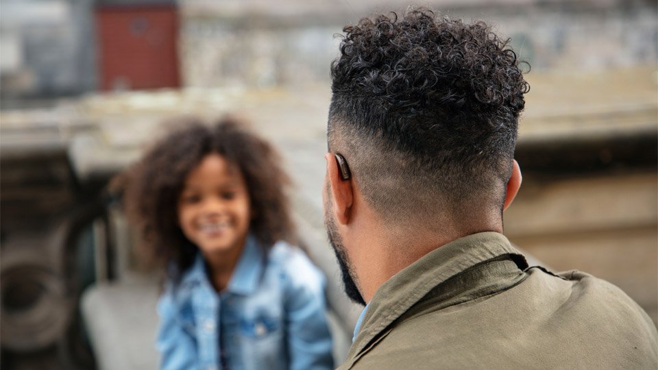 Man with short curly hair wearing a digital hearing aid, sitting outdoors and facing a young girl in a blurred background.