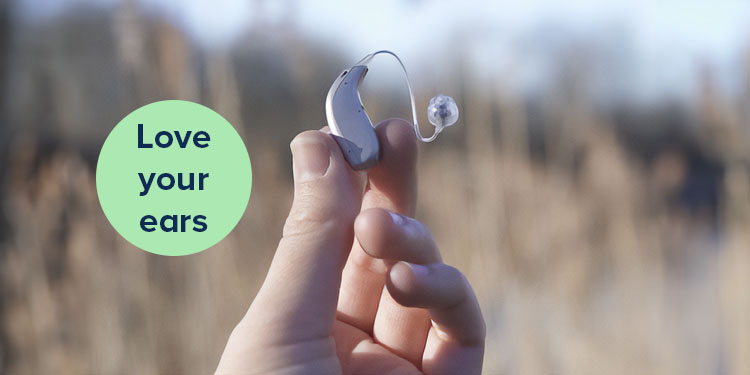 A hand holds a sleek modern hearing aid outdoors against a blurred natural background. Text reads: 'Love your ears' in a light green circle.
