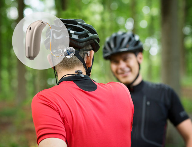 A cyclist wearing a black helmet and red jersey is engaged in conversation with another cyclist in a forest setting. A circular overlay highlights an Oticon hearing aid discreetly positioned behind their ear, emphasizing advanced hearing care solutions for active lifestyles.