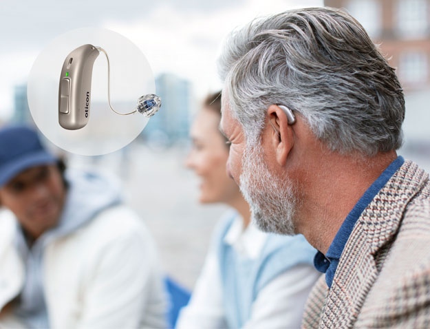 A gray-haired man converses with blurred individuals outdoors near a waterfront; an Oticon hearing aid is highlighted in an inset circle, showcasing advanced hearing aid technology for clear sound.