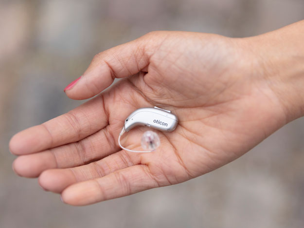 A hand holds a sleek Oticon hearing aid with a clear earpiece, positioned against a blurred neutral background, highlighting modern hearing technology provided by a hearing care specialist.