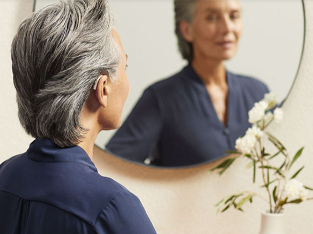 Eine grauhaarige Person trägt eine dunkelblaue Bluse, betrachtet sich in einem runden Spiegel und steht in einem hell eingerichteten Raum mit einer Vase weisser Blumen auf einem Tisch.