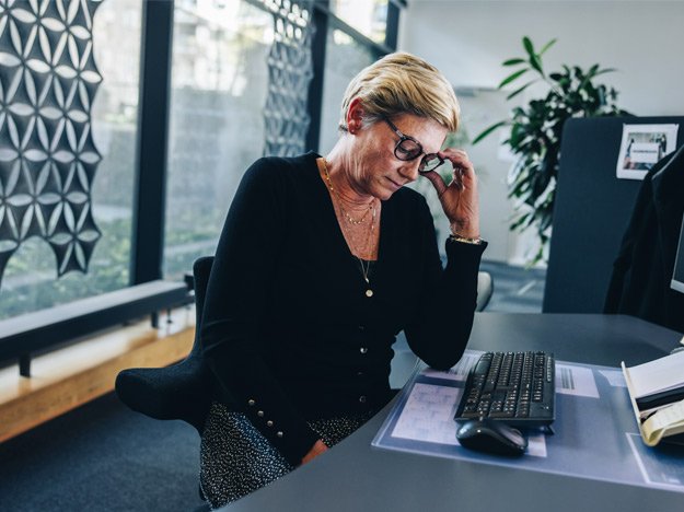 Woman at a desk holding her ear, appearing distressed, potentially due to untreated hearing loss.