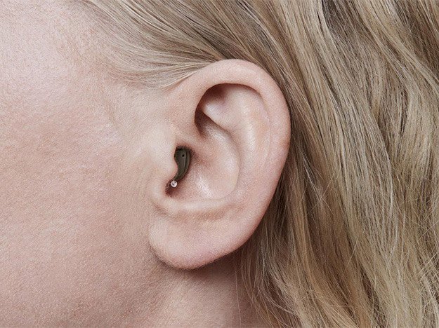 The cropped image features a close-up of a human ear surrounded by light blond hair, with part of the face blurred. It appears in a hearing care-related context.