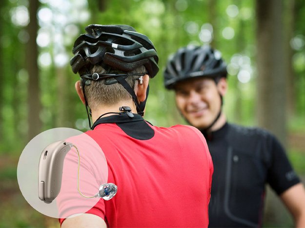 A cyclist wearing a red jersey and black helmet, equipped with a hearing aid, interacts with another cyclist in a forested area. A detailed inset highlights the hearing aid design.