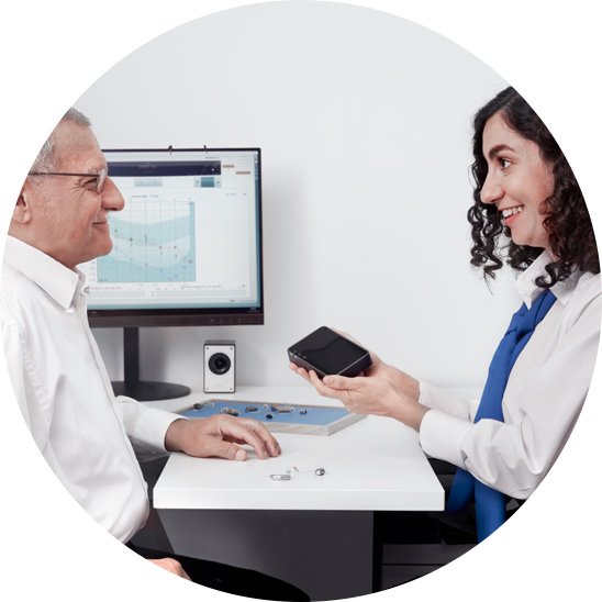 Audiologist demonstrating hearing aid options to a customer during a consultation, with an audiogram displayed on a monitor in the background.