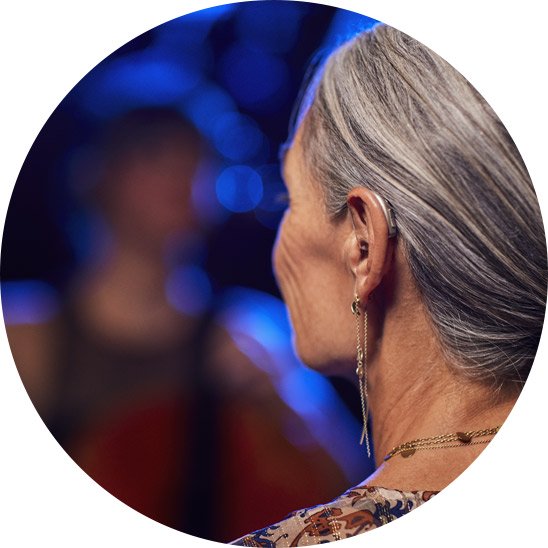 Woman with gray hair wearing a digital hearing aid, attending an event with blurred lights and a person in the background.