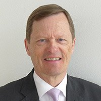 Un homme en costume sombre et cravate rose clair se tient devant un fond uni gris clair. Son visage est flouté, rendant impossible l’identification de ses traits ou expressions.