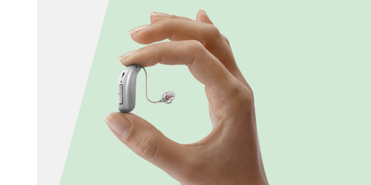 A hand delicately holds a sleek behind-the-ear hearing aid with a slim, transparent tube and earpiece against a light green background, emphasizing advanced hearing care technology and design.