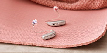 Deux appareils auditifs argentés à contour d’oreille, dotés d’embouts colorés (rouge et bleu), reposent sur une surface rose texturée, près d’un tissu discret.