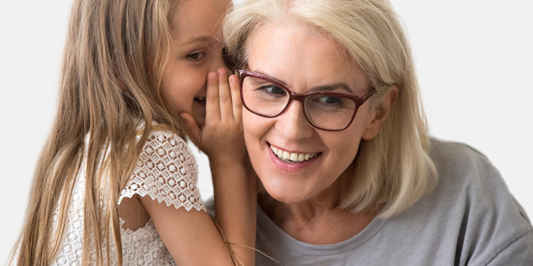A young girl leans in to whisper into an older woman's ear in a neutral, light-colored setting. The image emphasizes the importance of hearing, such as through hearing care services like tests or aids. No text is present.