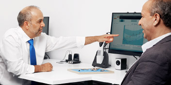 Un audiologiste, vêtu d’une chemise blanche et cravate bleue, pointe un écran affichant un audiogramme. Un patient l’écoute dans une salle de consultation du centre Audika.