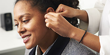 A hearing care specialist adjusts a hearing aid behind a patient's ear in a professional clinic setting, with a speaker and desk visible in the background.