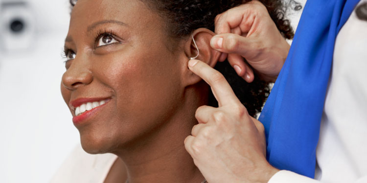 Un audioprothésiste ajuste un appareil auditif discret derrière l’oreille d’un patient, dans un contexte clinique lumineux.