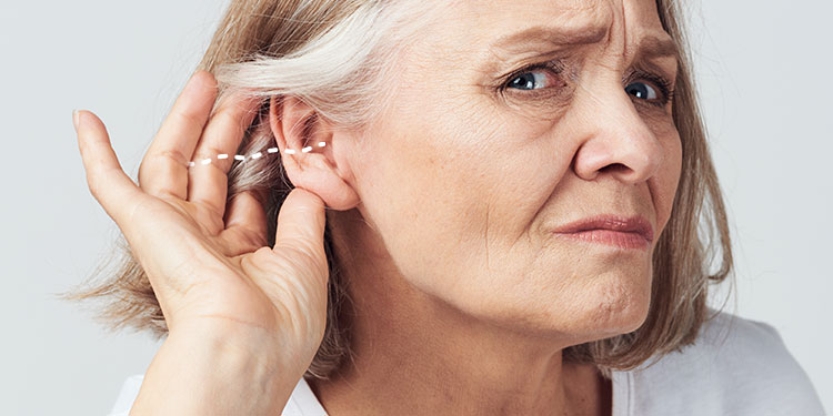 [GEN] A person's hand adjusts a discreet hearing aid positioned behind their ear, with dotted lines emphasizing placement; the setting appears to be a clinical or promotional context for hearing care. No text is visible.