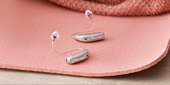 Deux appareils auditifs argentés à contour d’oreille, dotés d’embouts colorés (rouge et bleu), reposent sur une surface rose texturée, près d’un tissu discret.