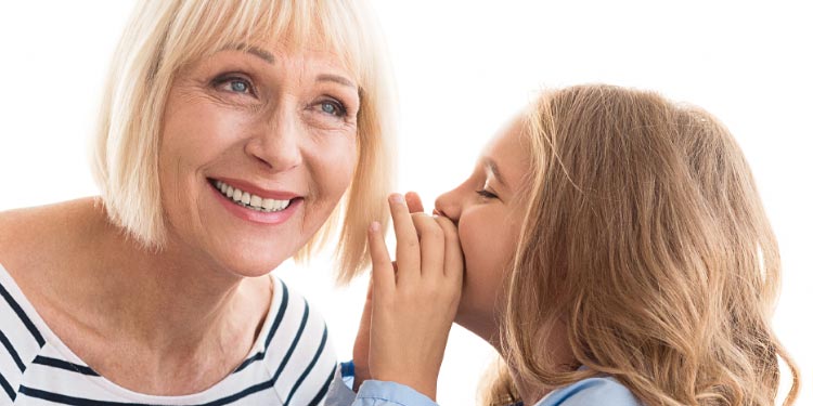Uma mulher mais velha, vestindo uma blusa listrada, escuta atentamente enquanto uma jovem com cabelos castanhos e camisa azul sussurra algo em seu ouvido. Fundo claro e neutro.