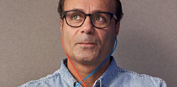 Una persona con rostro desenfocado lleva un audiómetro de tubos azul y rojo colgado del cuello, vistiendo una camisa de mezclilla, en un entorno neutro de clínica auditiva.