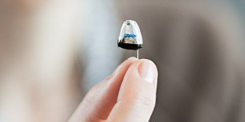 A hand holds a small, transparent in-ear hearing aid with detailed components visible inside. The background is softly blurred, suggesting a clinical or professional hearing care setting.