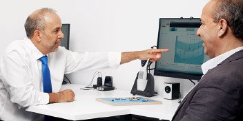 Un audiologiste, vêtu d’une chemise blanche et cravate bleue, pointe un écran affichant un audiogramme. Un patient l’écoute dans une salle de consultation du centre Audika.