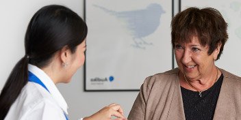 Een audioloog (met witte jas en blauw lint) toont een gehoorapparaat aan een oudere vrouw in een gehoorkliniek. Op de achtergrond hangt een Audika-poster met een vogelafbeelding.