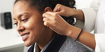Een audioloog past een hoortoestel aan bij een persoon in een gehoorcentrum. De witte jas en professionele omgeving, met een luidspreker op de achtergrond, benadrukken de klinische setting.
