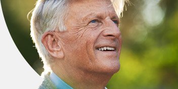 [GEN] A close-up of an older man's ear with gray hair, outdoors in a sunlit, green natural environment. No visible text or identifiable hearing aid brands are present in the image.