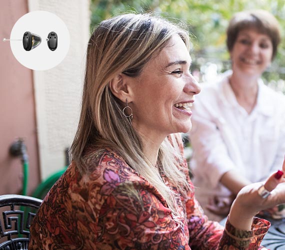Una donna con capelli chiari indossa un apparecchio acustico discreto nell’orecchio destro, conversando con un’altra persona in un ambiente esterno circondato da vegetazione. Nella foto appare un’immagine ingrandita di un apparecchio acustico.