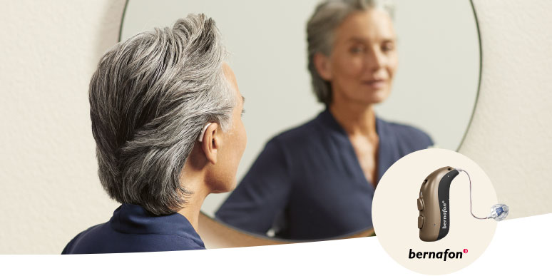 A person with short, silver-gray hair stands in front of a round mirror, wearing a navy-blue top. The image prominently features a Bernafon hearing aid, displayed separately in the corner with the brand logo for visibility.