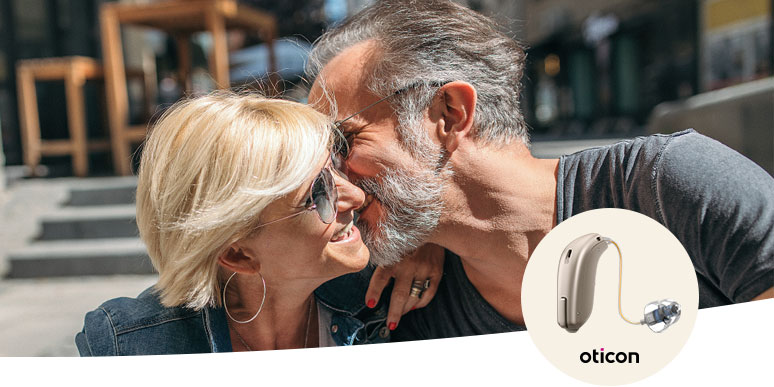 Um casal sentado em um ambiente urbano, possivelmente ao ar livre, em um momento de afeto. No canto inferior direito, destaca-se a imagem de um aparelho auditivo da marca Oticon, associado a cuidados auditivos.