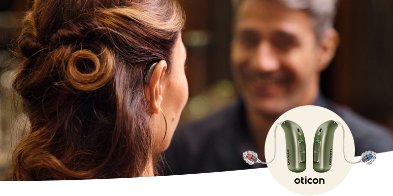 Een vrouw met opgestoken bruin haar luistert naar een persoon (gezicht onscherp) in een gesprek. In de hoek staan Oticon-hoortoestellen afgebeeld, relevant voor hoorzorg en hoorklinieken.