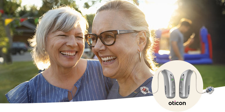 Twee vrouwen staan buiten in een zonnig park met feestelijke kleurrijke vlaggetjes op de achtergrond. Rechts onderaan worden Oticon hoortoestellen getoond, ideaal voor gehoorzorg bij Audika.
