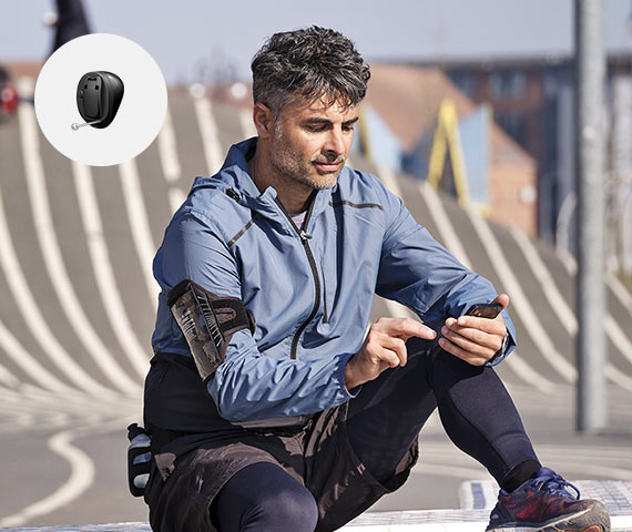 A man in athletic attire sits outdoors on an urban pathway, using a smartphone. A circular insert displays an Oticon hearing aid, highlighting advanced hearing solutions for active lifestyles.