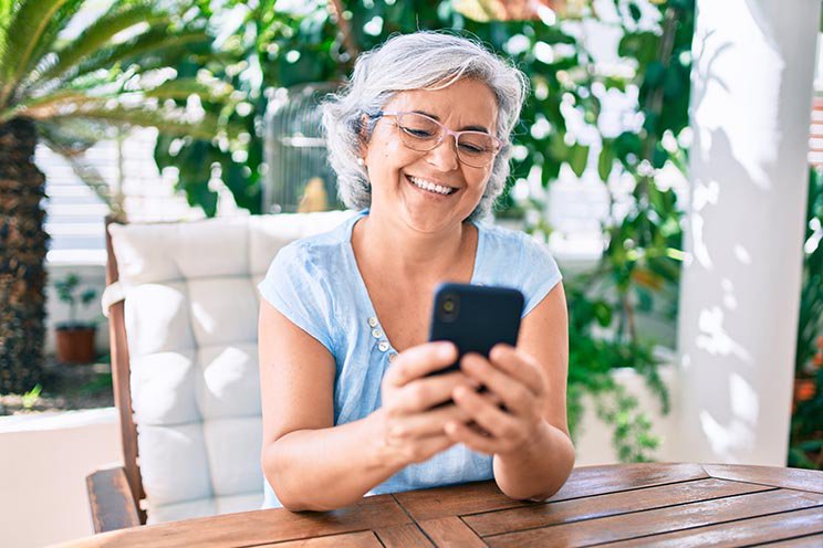 Mujer mayor usando un teléfono móvil mientras se sienta al aire libre en un entorno luminoso y natural.