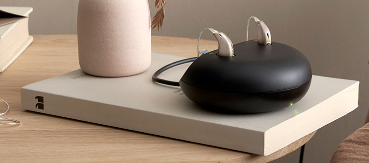 Deux appareils auditifs beige doré posés sur une station de recharge noire, disposée sur un livre posé sur une table en bois.