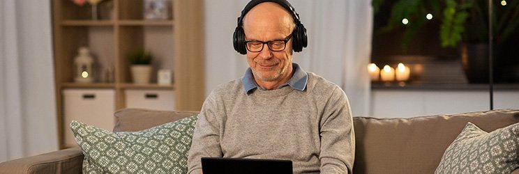 Man wearing headphones, sitting on a sofa with a laptop, in a cozy living room setting.