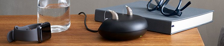 Two hearing aids rest on a sleek black charging station, placed on a wooden table alongside a smartwatch, a glass of water, eyeglasses, and a hardcover book.