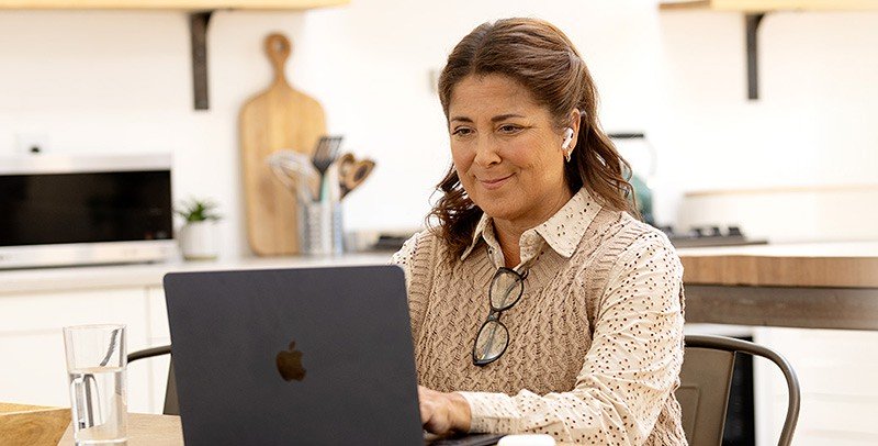 Mulher a usar aparelho auditivo e trabalhar em um notebook Apple, sentada em uma cozinha moderna com utensílios e eletrodomésticos ao fundo. Um copo de água está próximo na mesa.