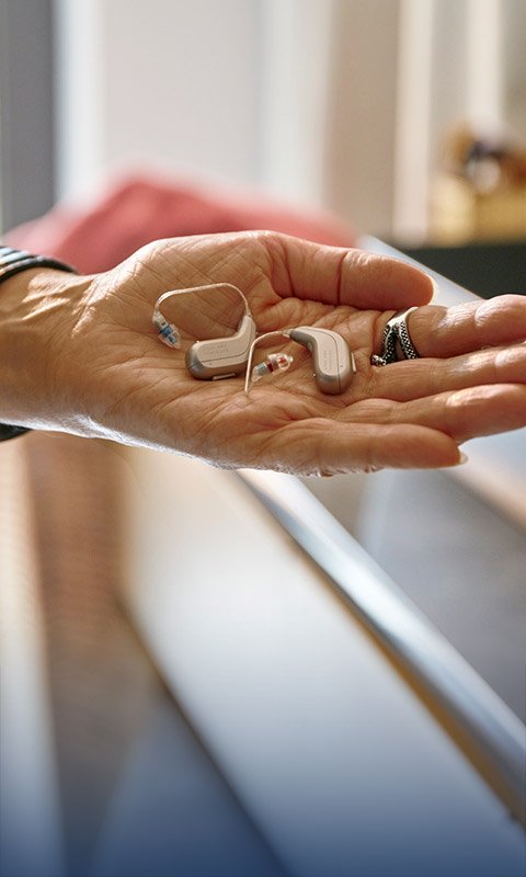 A hand holds two sleek hearing aids, likely from Oticon, featuring discreet design and color-coded tips, against a softly lit clinic environment with blurred background elements.