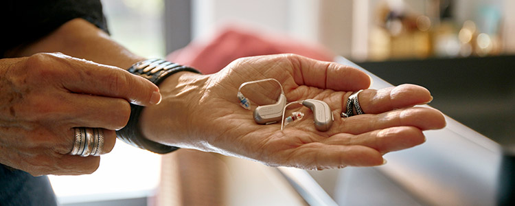 A hand holds three modern hearing aids, including in-the-ear and behind-the-ear models, in a bright and cozy interior setting, suggesting professional hearing care at a clinic or home.