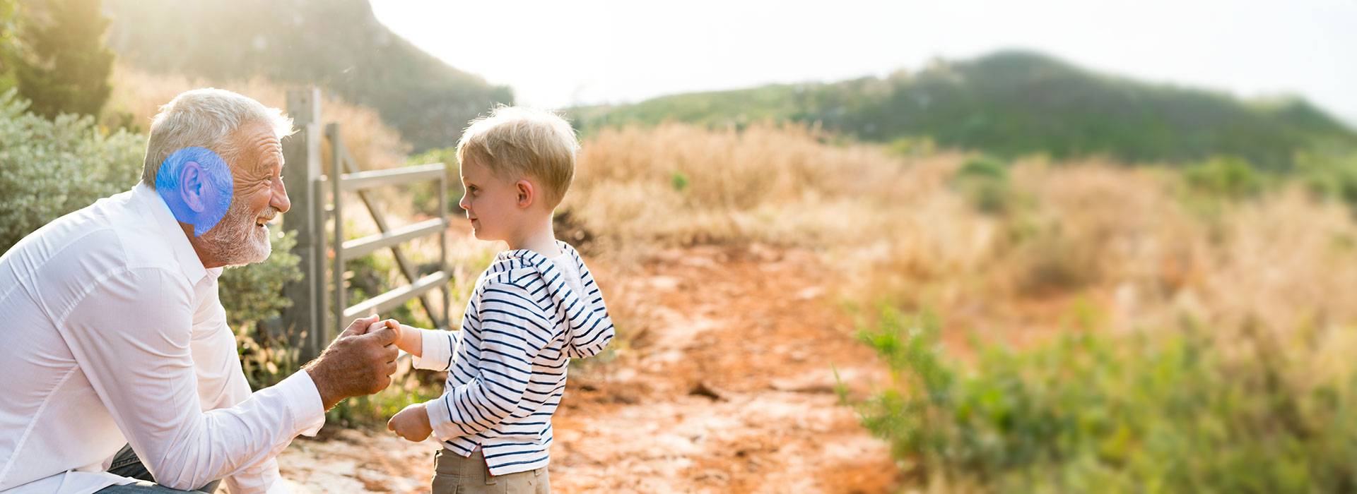 Un uomo anziano, con indosso un apparecchio acustico, si china e interagisce con un bambino in un sentiero all’aperto, circondato da vegetazione e colline luminose.