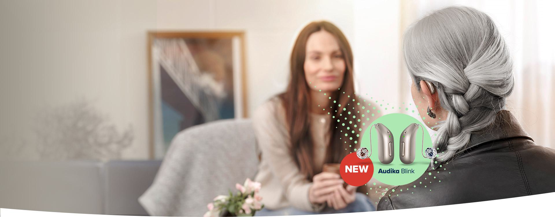 Una donna anziana con capelli grigi intrecciati indossa un apparecchio acustico in una sala luminosa, mentre parla con una donna giovane sfocata. Testo visibile: ’NEW Audika Blink’.