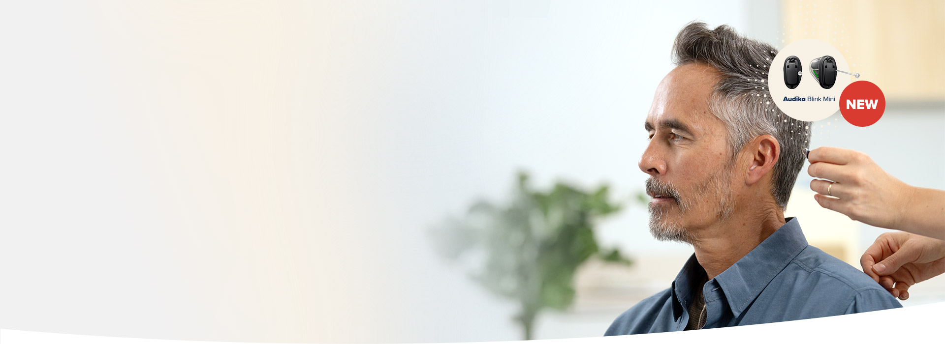 Una mano posiziona un apparecchio acustico 'Audika Blink Mini NEW' dietro l'orecchio di un uomo seduto in un centro acustico, con sfondo sfocato e pianta verde.