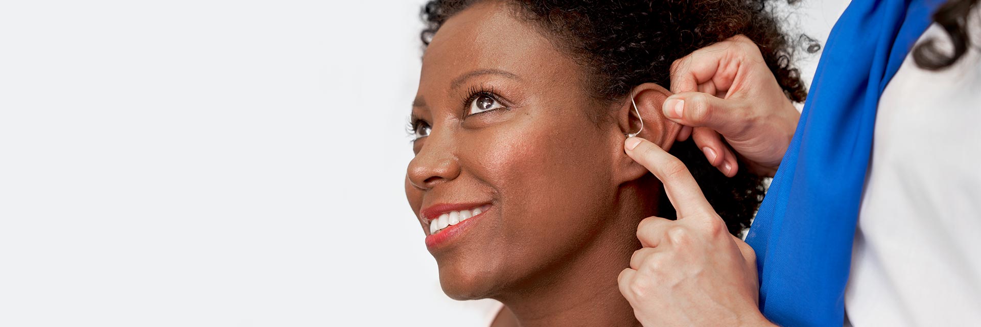 A hearing aid is being adjusted behind a person's ear by an audiologist wearing a blue scarf, in a clinical setting associated with hearing care services. No visible text or branding present.