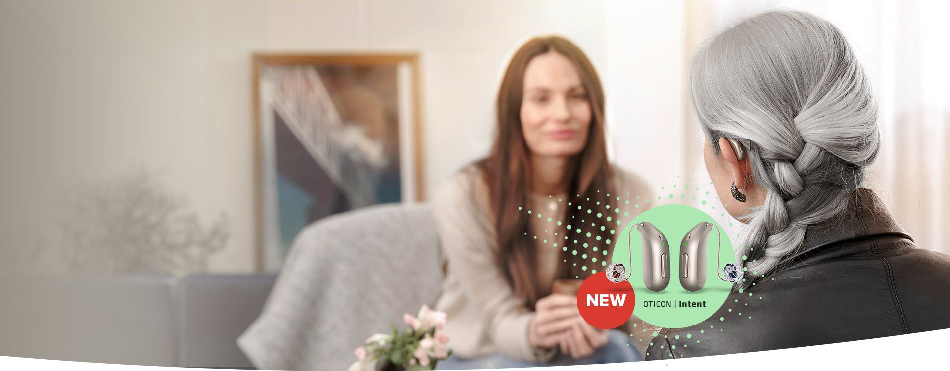 Une femme aux cheveux gris porte un appareil auditif Oticon. Elle discute avec une autre femme floue dans un environnement de salon. Texte : « NEW OTICON | Intent ».