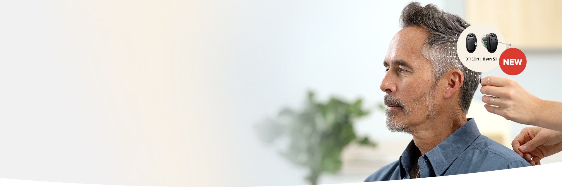 A clinician fits a man with an Oticon Own S1 hearing aid, highlighted with a 'NEW' label. The setting is a bright, professional hearing clinic with a blurred plant in the background.