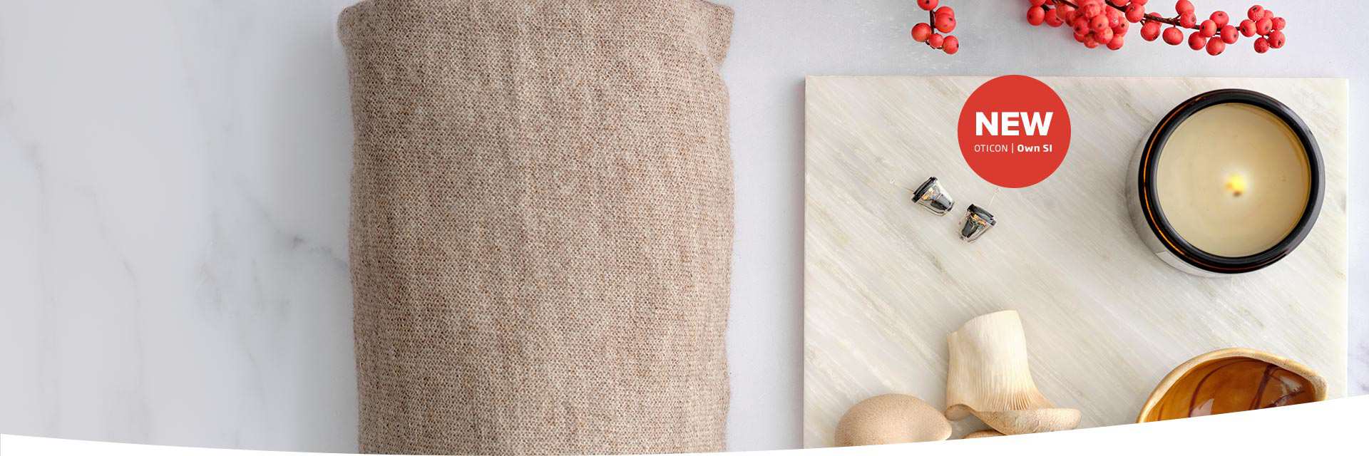 Two Oticon Own S1 in-the-ear hearing aids are displayed on a marble surface alongside a lit candle, wooden decor, red berries, and a beige woven fabric. Text reads: 'NEW OTICON | Own S1.'