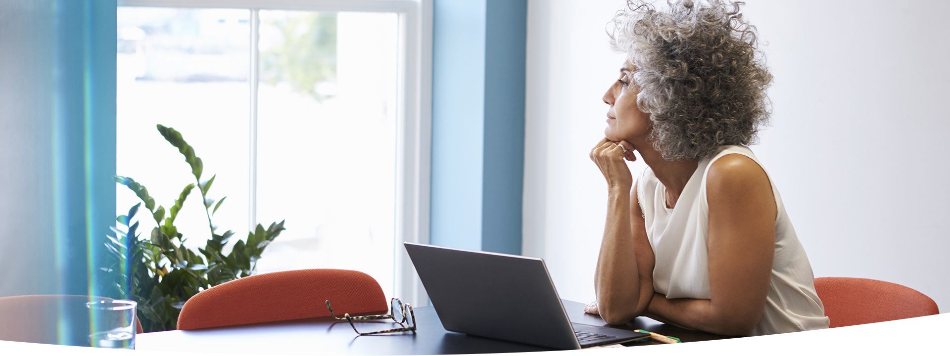A woman with curly gray hair, seated at a table in a brightly lit room, rests her chin on her hand, with a laptop, glasses, and a glass of water nearby.